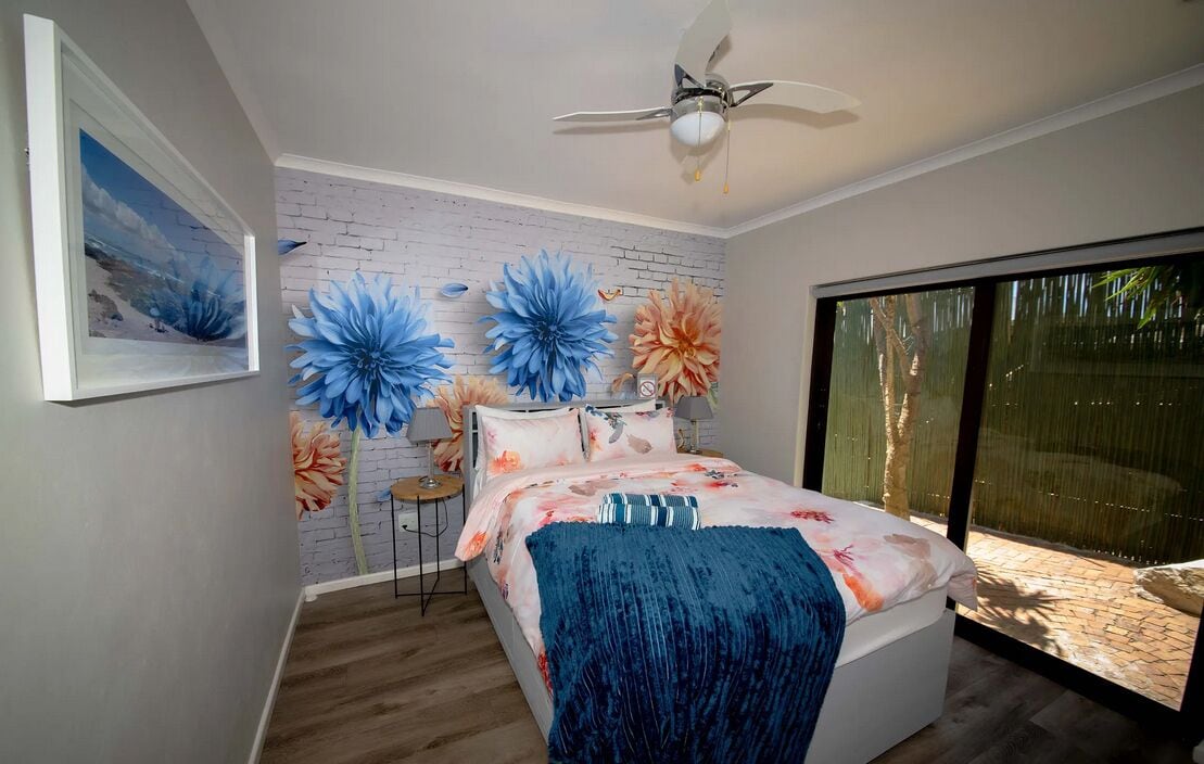 A bedroom with a bed, a ceiling fan, a large window, and a brick wall.