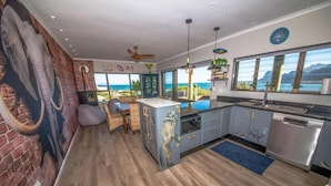 A modern kitchen with a large island, a dining area with a view, and a brick accent wall. - The Butterfly Villa (Pringle Bay)