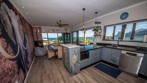 A modern kitchen with a view of the ocean, a brick wall with a large elephant sculpture, and a dining area with wicker furniture. - The Butterfly Villa (Pringle Bay)