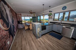 Une cuisine moderne avec vue sur l’océan, un mur en briques orné d’une grande sculpture d’éléphant et un coin repas avec des meubles en osier.