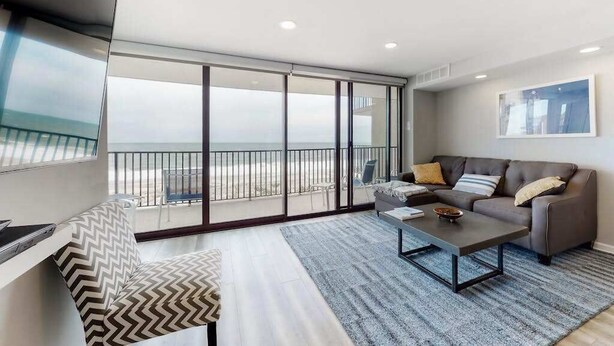 Una sala de estar moderna con un sofá, una mesa de centro, un televisor y grandes puertas correderas de vidrio que dan a un balcón con vistas a la playa.