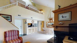 A spacious living area with a high ceiling, a fireplace, a dining area with chairs, and a staircase leading to an upper level. - Sea Colony Tennis 2nd floor condo w/ gym, shared sauna, and balcony (Bethany Beach)