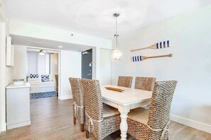 Un coin repas avec une table blanche et des chaises en osier, un luminaire suspendu et une vue sur une autre pièce.