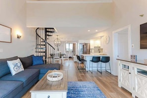 A modern living room with a sofa, coffee table, and a staircase leading to a kitchen area. - Sea Colony oceanfront penthouse w/basketball court, hot tub, pool, tennis & more (Bethany Beach)