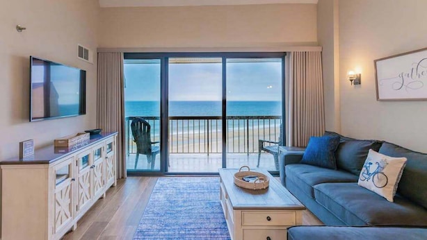 Un soggiorno con televisione, un divano, un tavolino da caffè e vista sulla spiaggia attraverso ampie porte scorrevoli.