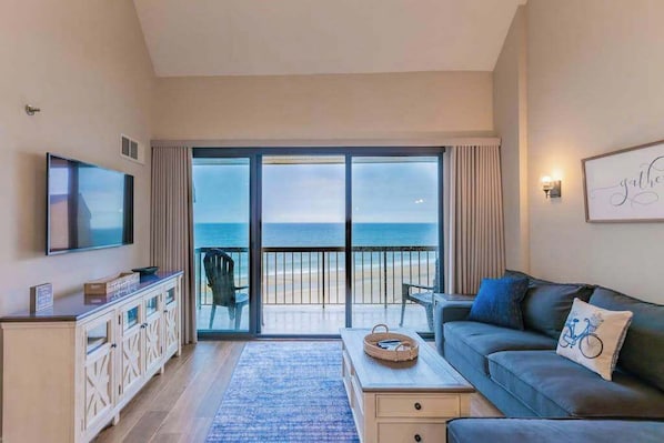A living room with a television, a sofa, a coffee table, and a view of the beach through large sliding doors. - Sea Colony oceanfront penthouse w/basketball court, hot tub, pool, tennis & more (Bethany Beach)