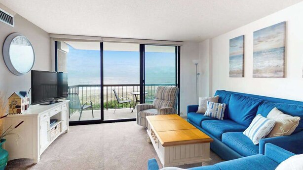 A living room with a blue sofa, a wooden coffee table, a TV on a white cabinet, and a balcony with a view of the ocean. - Sea Colony Ocean 3rd floor condo w/ pool, tennis court, and elevator (Bethany Beach)