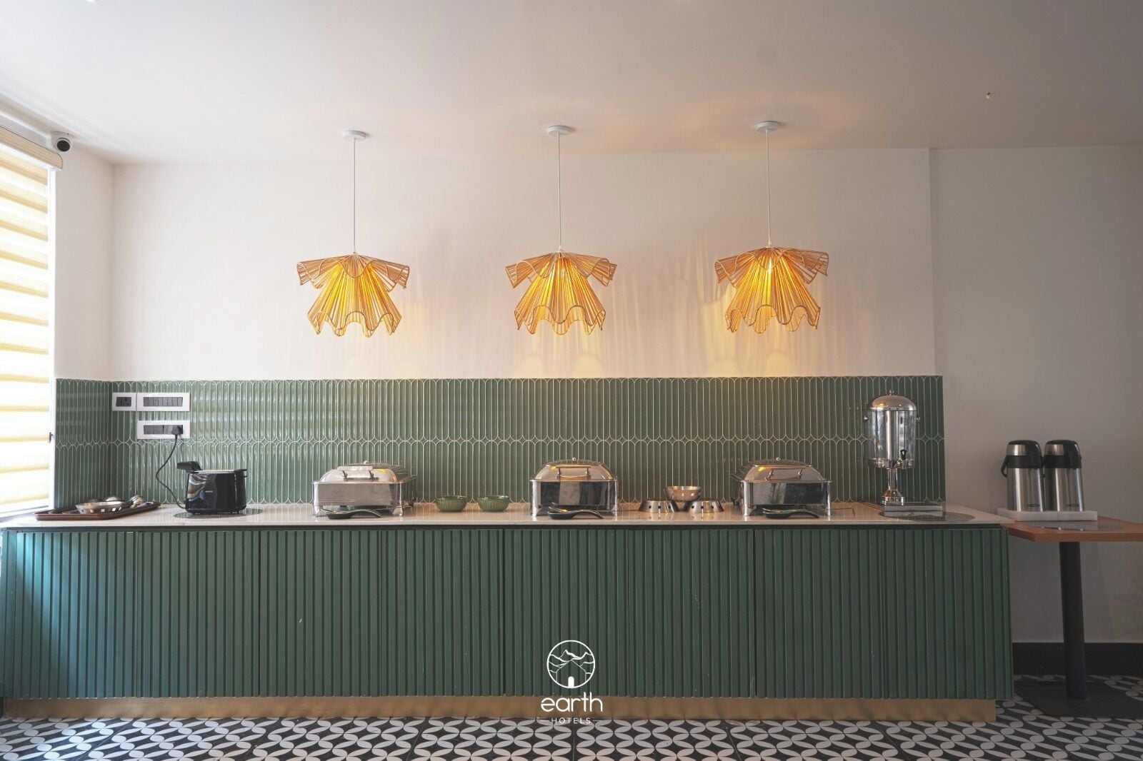 Cozinha de hotel com bancada verde, três luminárias pendentes e diversos eletrodomésticos.