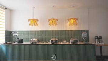 Cozinha de hotel com bancada verde, três luminárias pendentes e diversos eletrodomésticos.