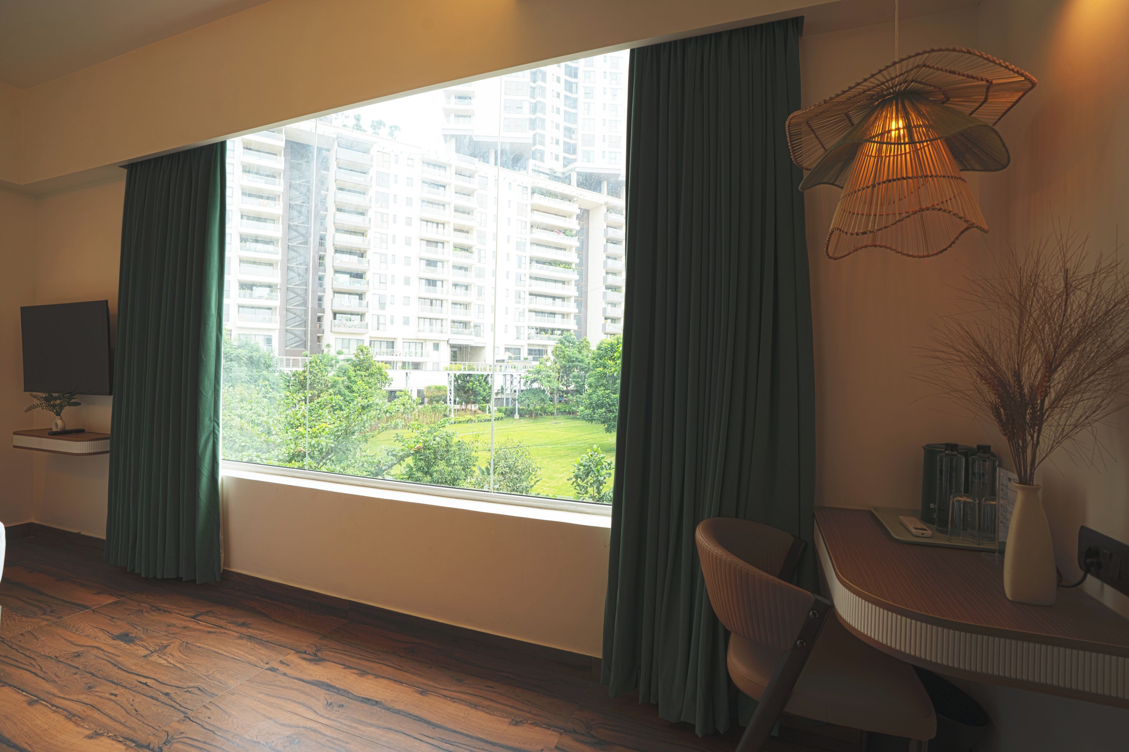 A hotel room with a large window, a desk with a chair, a television, and a decorative hanging light fixture.