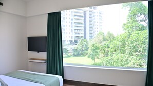 A hotel room with a bed, a flat-screen TV mounted on the wall, a window with curtains, and a view of a building and greenery outside. - Earth Hotels Hebbal (Bengaluru)
