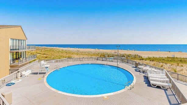 A pool with a view of the beach and ocean, surrounded by lounge chairs and a building with balconies. - Sea Colony Ocean 6th-floor condo wit balcony, tennis court, & basketball court (Bethany Beach)
