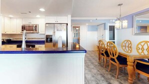A modern kitchen with white cabinets, a blue countertop, stainless steel appliances, and a dining area with wooden chairs and a table. - Sea Colony Ocean 6th-floor condo wit balcony, tennis court, & basketball court (Bethany Beach)