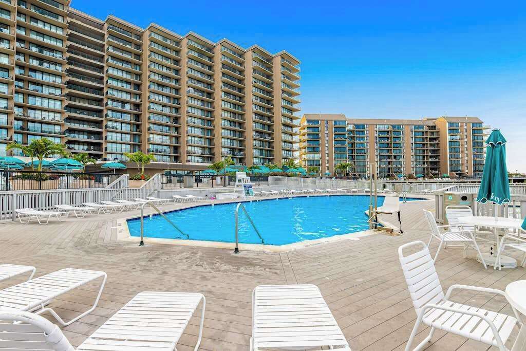 A hotel pool area with multiple lounge chairs, a swimming pool, and high-rise buildings in the background.