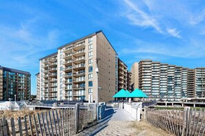 Un edificio residencial de varios pisos con balcones, una galería techada y una zona arenosa al frente.