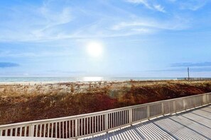 Vista a la playa desde una terraza de madera con barandilla, cielo despejado y el océano al fondo.