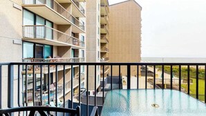 Blick auf einen Pool mit Glasbarriere, ein Gebäude mit mehreren Balkonen und freier Sicht auf den Strand.