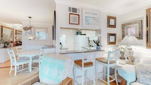 Un comedor con mobiliario blanco, una cocina al fondo y cuadros en las paredes.