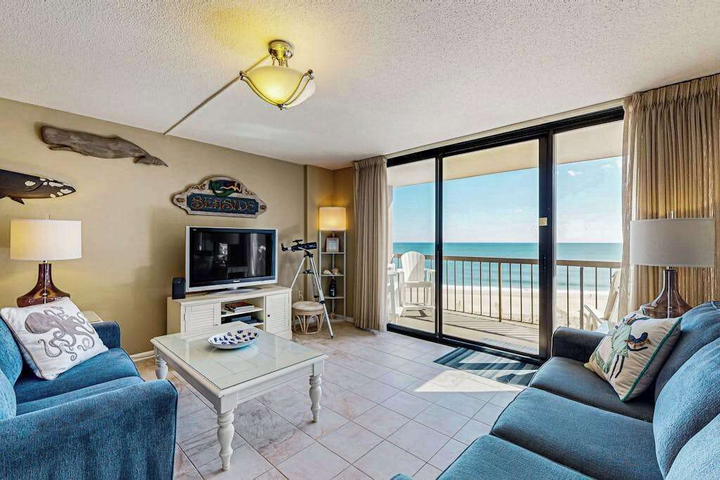 Ein Wohnzimmer mit Fernseher, Stativ und Balkon mit Blick auf den Strand.