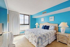 A bedroom with a large bed, two bedside tables with lamps, a window with curtains, and a view of the sky. - Oceanfront fifth-floor condo with sauna, pools, & hot tub on-site (Bethany Beach)