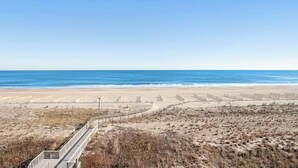 Une plage avec une promenade en bord de mer qui mène à l’océan, des dunes de sable et un ciel dégagé.