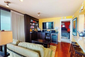 A compact living space with a sofa, a TV, a bookshelf, and a kitchenette. - Sea Colony 3rd floor condo w/ shared sauna, gym, pool & ocean view (Bethany Beach)