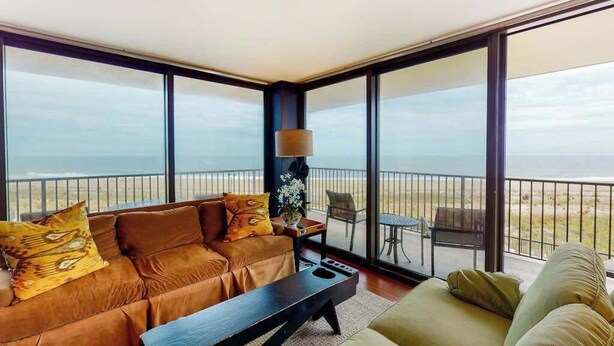 A modern living room with a large window, a sofa, a coffee table, and a small table with a lamp. - Sea Colony 3rd floor condo w/ shared sauna, gym, pool & ocean view (Bethany Beach)
