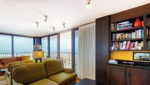 A modern living room with a sofa, a coffee table, a bookshelf, and a view of the ocean through large windows. - Sea Colony 3rd floor condo w/ shared sauna, gym, pool & ocean view (Bethany Beach)