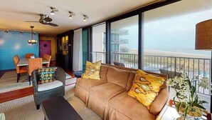 A modern living room with a sofa, a dining table, and chairs. There is a balcony with a railing and a view of the outside. - Sea Colony 3rd floor condo w/ shared sauna, gym, pool & ocean view (Bethany Beach)