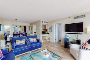 A living room with a blue sofa, a white sofa, a glass coffee table, and a flat-screen TV mounted on the wall. - Sea Colony Ocean 1st floor condo w/ tennis court, balcony, and pool (Bethany Beach)