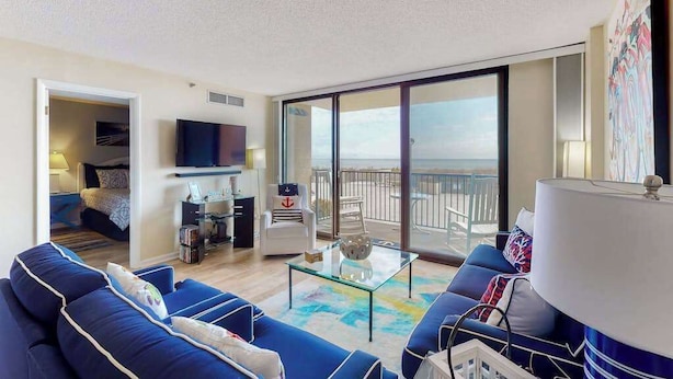 Una sala de estar moderna con sofá, una mesa de centro y televisor. Hay un balcón con vistas al mar.