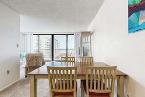 Un espace repas avec une table et des chaises en bois, une vue sur les bâtiments à travers de grandes fenêtres et un tableau accroché au mur.