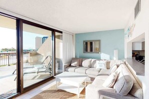 A modern living room with a sectional sofa, a glass sliding door leading to a balcony, and a minimalist decor. - Sea Colony Ocean 1st floor condo w/ basketball court, tennis court, & WiFi (Bethany Beach)