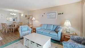 Ein Wohnzimmer mit einem blauen Sofa, einem weißen Couchtisch, Korbstühlen, einem Esstisch und einer Lampe.