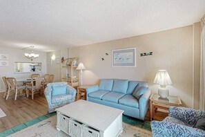 A living room with a blue sofa, a white coffee table, wicker chairs, a dining table, and a lamp. - Sea Colony 8th floor condo w/ shared gym, sauna, basketball court - ocean view (Bethany Beach)