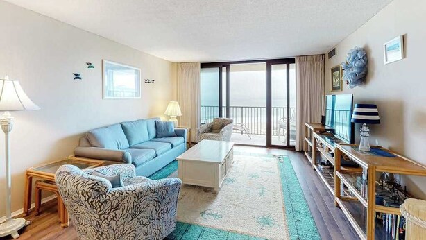 Ein geräumiges Wohnzimmer mit einem blauen Sofa, einem gemusterten Sessel und einem Couchtisch. Ein Bücherregal und ein großes Fenster mit Blick sind ebenfalls vorhanden.