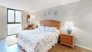 Ein Schlafzimmer mit einem großen Bett, einem Holz-Kopfteil, Nachttischlampen, einem Nachttisch und Blick durch ein großes Fenster.