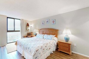 A bedroom with a large bed, wooden headboard, bedside lamps, a nightstand, and a view through a large window. - Sea Colony 8th floor condo w/ shared gym, sauna, basketball court - ocean view (Bethany Beach)