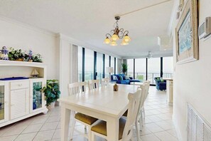 Een ruime kamer met een eettafel, stoelen, een kroonluchter, een bank en grote ramen.