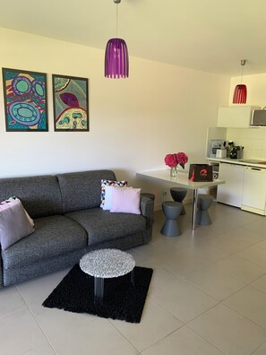 Uma sala de estar moderna com um sofá cinzento, uma mesinha redonda e um tapete preto. Há quadros abstratos na parede, uma luminária pendente roxa e uma luminária suspensa vermelha. Uma área de cozinha é visível ao fundo.