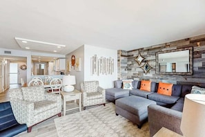 A modern living room with a sectional sofa, patterned armchairs, a dining area, and a kitchen in the background. - Sea Colony Ocean 14th floor condo with elevator, WiFi, and basketball court (Bethany Beach)