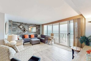 En rummelig stue med en stor sofasæt, et sofabord og et spiseområde. Der er en balkon med havudsigt.