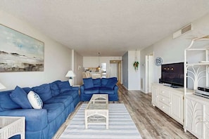 Un salon avec un canapé bleu, une table basse, un téléviseur posé sur un meuble blanc et un tableau accroché au mur.
