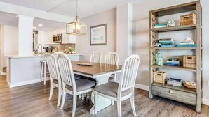A dining area with a wooden table and white chairs, a kitchen in the background, and a bookshelf with various items. - Sea Colony 1st floor condo w/ tennis court, shared sauna & balcony w/ ocean view (Bethany Beach)