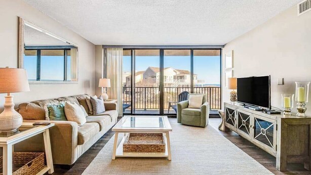 A living room with a sofa, armchair, TV, and a balcony with a view. - Sea Colony 1st floor condo w/ tennis court, shared sauna & balcony w/ ocean view (Bethany Beach)