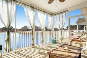 A covered deck with a view of a lake, a table with a shell, and lounge chairs.