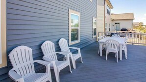 Eine Terrasse mit weißen Adirondack-Stühlen und einem Tisch, ein Haus mit blauen Wandverkleidungen und einem wolkenlosen Himmel.