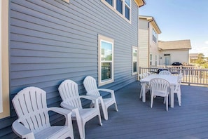 Eine Terrasse mit weißen Adirondack-Stühlen und einem Tisch, ein Haus mit blauen Wandverkleidungen und einem wolkenlosen Himmel.
