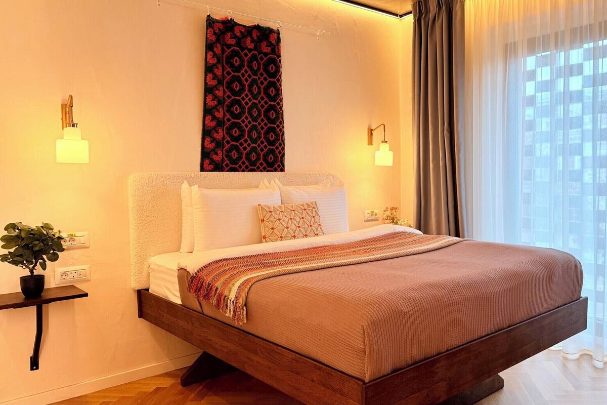 A modern hotel room with a large bed, bedside lamps, a wall-mounted tapestry, and a window with sheer curtains.