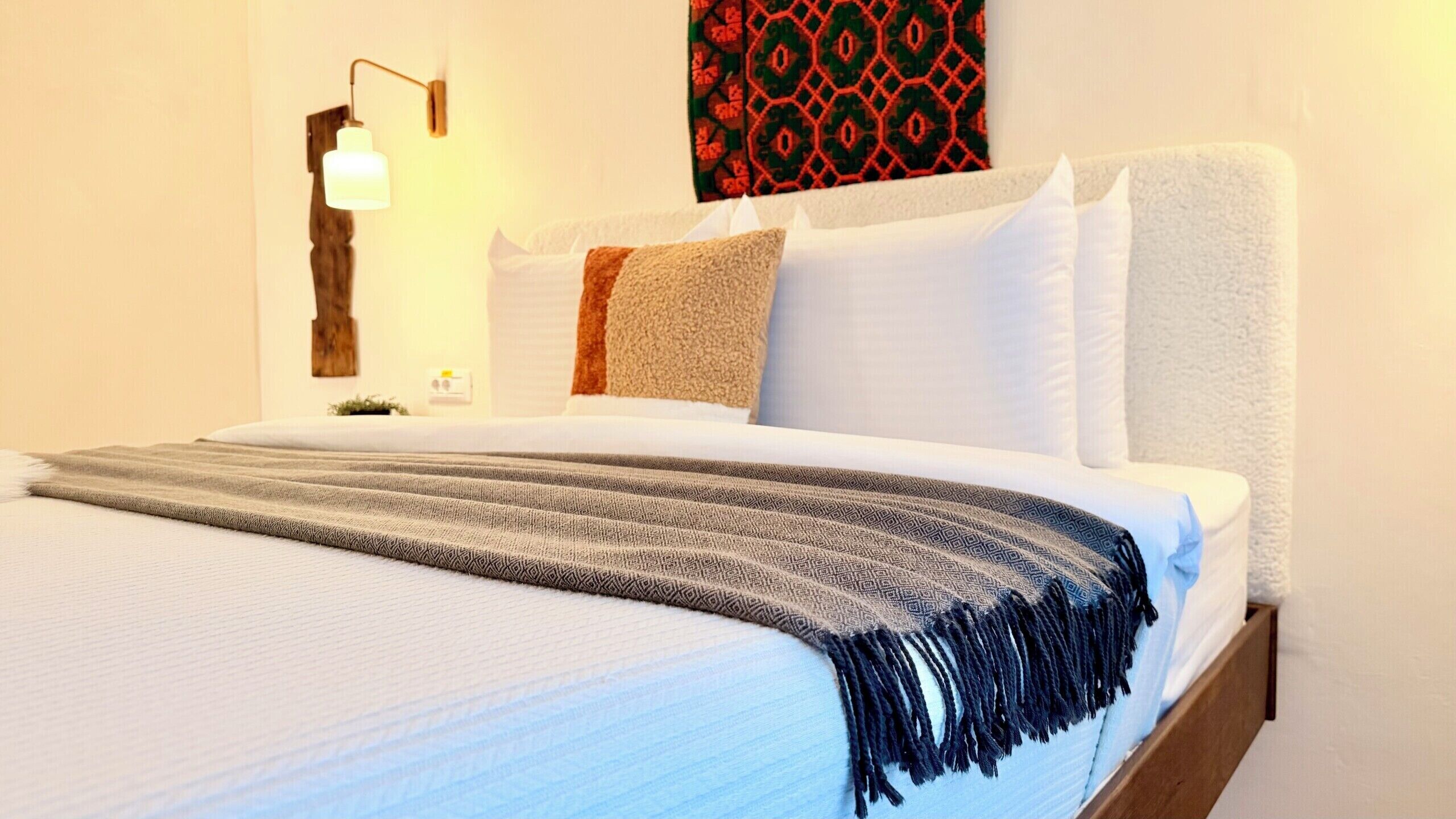 Cama bem arrumada com manta estampada, luminária fixada na parede e um enfeite de parede decorativo.